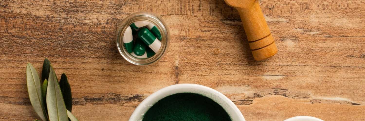 Un bol contenant de la spiruline en poudre accompagné de comprimés de spiruline dans un bocal en verre. Les éléments sont sur une table en bois.