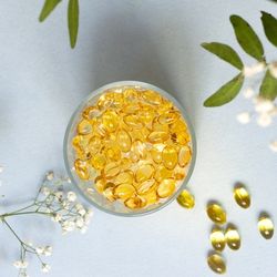 Capsules dorées d'acides gras essentiels dans un récipient en verre, entourées de feuilles et de petites fleurs blanches, symbolisant leur rôle dans une alimentation équilibrée et la santé.