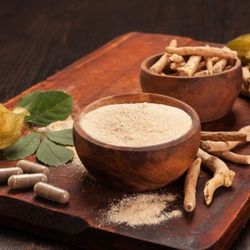 Bol en bois rempli de poudre d’ashwagandha et un autre bol contenant des racines séchées, avec des capsules et des feuilles vertes sur une planche en bois sombre.