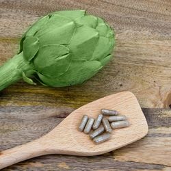 Capsules posées sur une cuillère en bois avec un artichaut frais en arrière-plan, sur une table en bois.