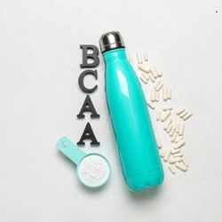 Une bouteille turquoise en métal, accompagnée de gélules blanches, d'une cuillère contenant de la poudre blanche et des lettres formant le mot 'BCAA', posées sur une surface claire.