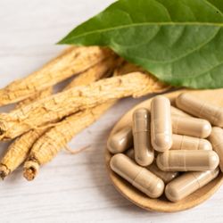 Racines de ginseng séchées et capsules beige clair sur une cuillère en bois, accompagnées de feuilles vertes fraîches.