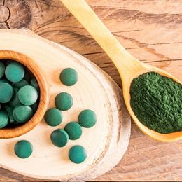 Photo de cachets de spiruline dans un bol en bois, posé à côté d'une cuillère en bois remplie de spiruline en poudre