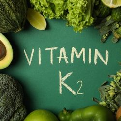 L'inscription "Vitamin K2" écrite sur une surface verte, entourée de légumes verts riches en cette vitamine, comme du brocoli, de l'avocat, de la laitue, des poivrons verts, et d'autres feuilles vertes.