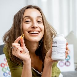 Une femme souriante tenant dans une main une boite de compléments alimentaires, et dans l'autre une gélule