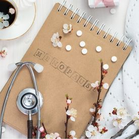 Carnet en spirale avec le mot 'Ménopause' écrit dessus, entouré de branches fleuries, de pilules blanches et d'un stéthoscope, posé sur une surface claire avec une tasse de café et un tissu blanc.