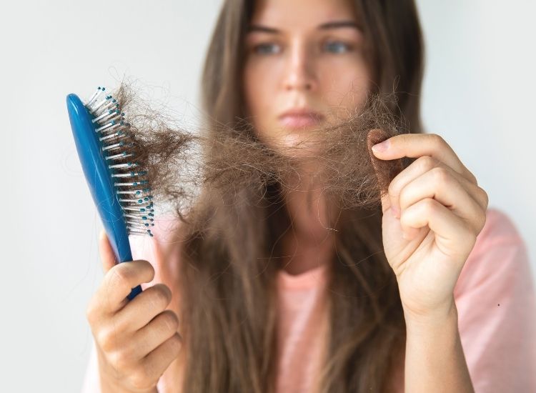 Photo d' une femme avec de longs cheveux bruns, tenant une brosse remplie de cheveux perdus. Elle semble préoccupée par la chute de ses cheveux.